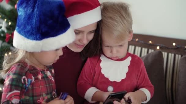 Noël, hiver, Nouvel An, Célébration, concept de famille heureux famille excitée vêtus de vêtements de Noël et santa chapeau faire l'achat en ligne tenir smartphone et carte de crédit assis près de l'arbre décoré. — Video