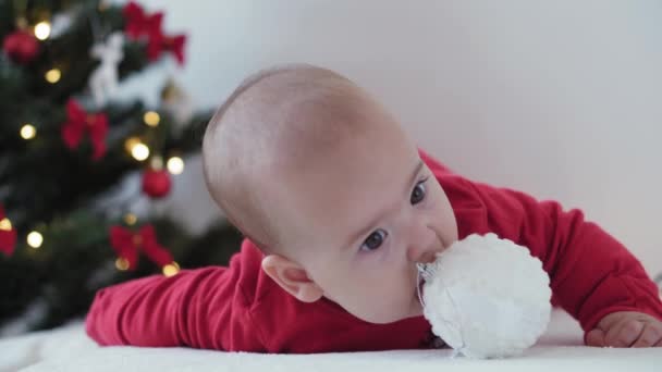 Feliz Natal e feliz ano novo, bebês, infância, conceito de férias - close-up bebê recém-nascido de 6 meses em chapéu de santa claus e bodysuit vermelho em sua barriga rasteja com bolas de decorações na árvore de natal — Vídeo de Stock