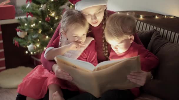 Kerstmis, winter, Nieuwjaar, Feestdag, Familie concept - Jonge gelukkige moeder twee kleine kinderen jongen en meisje gekleed in rode kleren gelezen sprookjes zitten in de buurt versierde kerstboom helder licht uit boek — Stockvideo