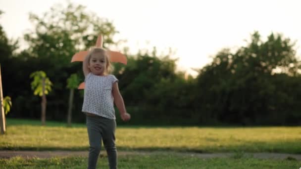 Infancia, juegos y entretenimiento, deportes, cultura física, parques y concepto al aire libre - niña pequeña rubia sosteniendo y lanzando avión de juguete naranja en el césped verde brillante en el parque de verano al atardecer. — Vídeos de Stock