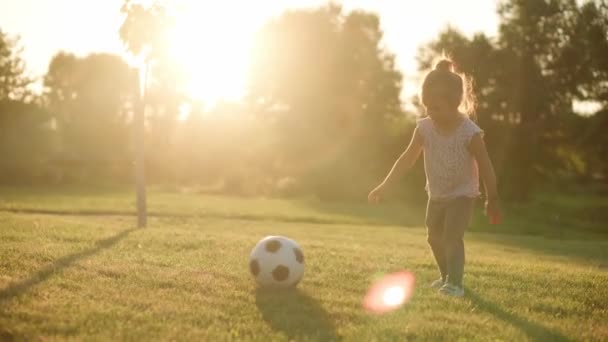 Infância, jogos e entretenimento, esportes, cultura física, parques e conceito ao ar livre - menina criança feliz com a jovem mãe jogar futebol com bola preta e branca no gramado verde no parque ao pôr do sol. — Vídeo de Stock