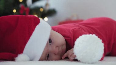 Mutlu Noeller ve mutlu yıllar, bebekler, çocukluk, tatil konsepti - yakın plan 6 aylık Noel Baba şapkalı bebek ve göbeğinde kırmızı tulum Noel ağacında süsleme toplarıyla sürünüyor