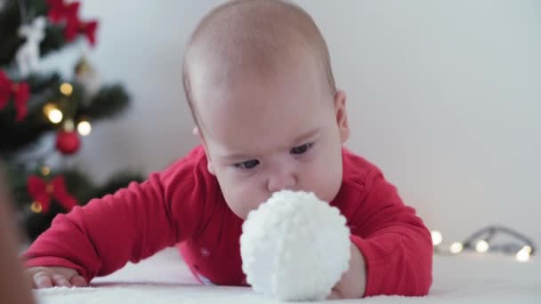 Feliz Navidad y feliz año nuevo, bebés, infancia, concepto de vacaciones - primer plano bebé recién nacido de 6 meses en sombrero de Santa Claus y traje de cuerpo rojo en su barriga se arrastra con bolas de decoración en el árbol de Navidad — Vídeo de stock