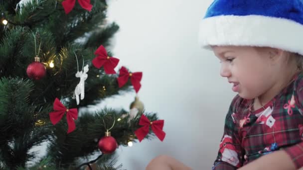 Natal, inverno, ano novo, celebração, conceito de família - menina sorrindo feliz vestida com roupas de Natal brinca com decorações, guirlandas e mostra emoção sentado perto da árvore decorada à noite — Vídeo de Stock