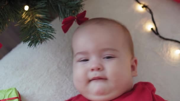Veselé Vánoce, šťastný nový rok, kojenci, dětství, dovolená koncept - close-up úsměv 6 měsíců staré novorozeně v Santa Claus klobouk a červené bodysuit ležel na zádech s dekoracemi v blízkosti vánočního stromku. — Stock video