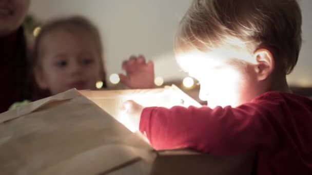 Noël, hiver, Nouvel An, Célébration, concept familial - Jeune maman heureuse avec deux petits enfants garçon et fille vêtus de vêtements rouges boîte cadeau ouverte assis près de l'arbre de Noël décoré. lumière vive de celui-ci — Video