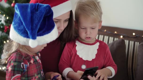 Vánoce, zima, Nový rok, Celebration, rodinný koncept - šťastný vzrušený rodina oblečená do vánočního oblečení a Santa klobouk, aby nákup on-line hold smartphone a kreditní karty sedí v blízkosti zdobeného stromu. — Stock video