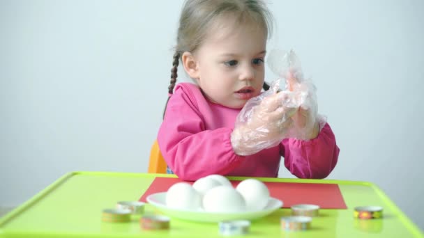 Pascua, Pascua, infancia, creatividad, concepto de juegos - primer plano de la pequeña rubia de tres años de edad, niña de aspecto caucásico esloveno en bata rosa pinta huevos de Pascua en guantes de plástico. — Vídeo de stock