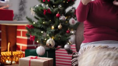 Xmas, kış, yeni yıl, kutlama, aile konsepti - Noel Baba şapkalı mutlu kız, karantinadaki Noel ağacının yanında oturan akıllı telefon dalgalarıyla tatildeki video konferansını uzaktan tebrik ediyor