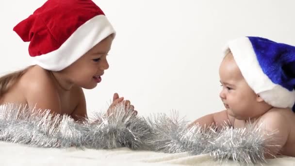 Весёлого Рождества и счастливого Нового года, младенец, детство, праздничная концепция - крупным планом 6-месячный новорожденный голый ребенок в красно-синей шляпе Санта-Клауса со старшей сестрой ползает с украшениями рождественской елки — стоковое видео