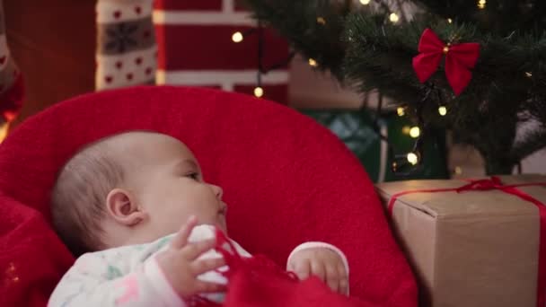 Xmas, winter, new year, Celebration, family concept - Beautiful smiling baby dressed in Christmas clothes sitting in rocking chair wraps in warm soft red blanket by the fireplace and Christmas tree. — Stock Video