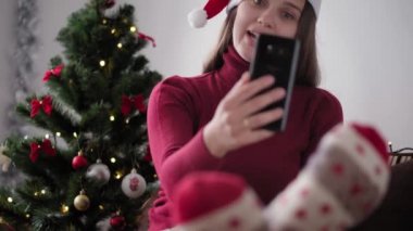 Xmas, kış, yeni yıl, kutlama, aile konsepti - Noel Baba şapkalı mutlu kız, karantinadaki Noel ağacının yanında oturan akıllı telefon dalgalarıyla tatildeki video konferansını uzaktan tebrik ediyor