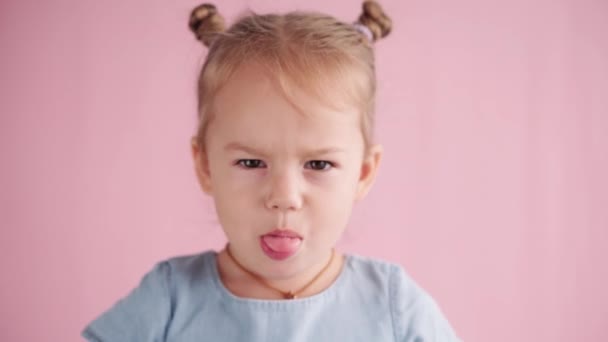 Детство, праздничная концепция - крупным планом счастливая блондинка в синем платье смотрит в камеру и показывает различные эмоции торчит язык и показывает недовольство на розовом твердом фоне. — стоковое видео