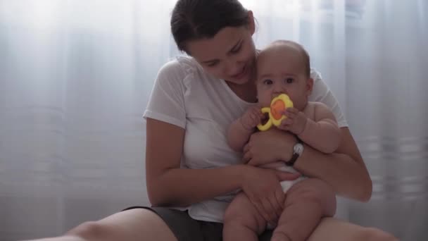 Medicine, healthcare, family, innocence, pediatrics, happiness, infant concepts - Young happy mother holding naked 4 month old baby and playing with colored toy tacroach sitting in nursery on floor — стоковое видео