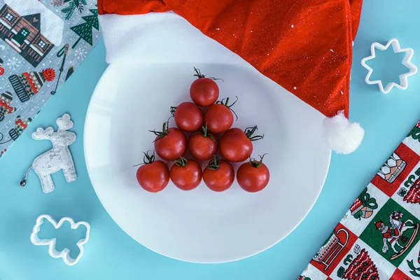 Natale, inverno, nuovo concetto anno - layout piatto bianco su cui ci sono albero di Natale foderato con pomodorini rossi ciliegio a forma di triangolo con cappello di Babbo Natale e prodotti da forno su sfondo blu — Foto Stock