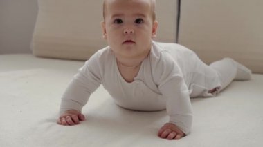 Tıp, sağlık, aile, masumiyet, pediatri, mutluluk, bebek konseptleri - Komik mutlu 7 aylık bebek karnının üstünde emekliyor ve parlak güneş ışığı kreşinde beyaz yumuşak yatakta sallanıyor.