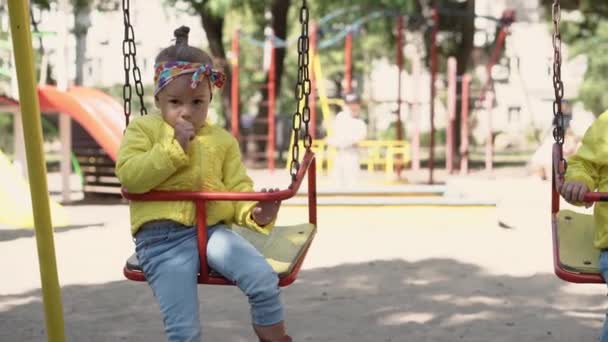 Childhood, game, entertainment, friendship, lifestyle concept - general plan of sad 3 year old girl Caucasian Slavic appearance in yellow jacket sit on swing pouting lips from disorder on playground. — Stock Video