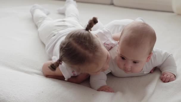 Medicína, zdravotní péče, rodina, nevinnost, pediatrie, štěstí, kojenecké pojmy - Funny happy 7 měsíců baby girl crawls on stomach with staršího sister girl in bright sunlight nursery on white soft bed. — Stock video