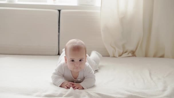 Medycyna, opieka zdrowotna, rodzina, niewinność, pediatria, szczęście, koncepcje niemowląt - Funny happy 7 miesięcy dziewczynka czołga się na brzuchu i kołysze się na kolanach w jasnym słońcu żłobek na białym miękkim łóżku. — Wideo stockowe