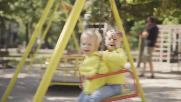 Infancia, juegos, entretenimiento, amistad, concepto de estilo de vida - plan general de dos niños en chaquetas amarillas niño y niña hermano y hermana 2 y 3 años de edad paseo alegremente en columpio en el patio de recreo — Vídeo de stock