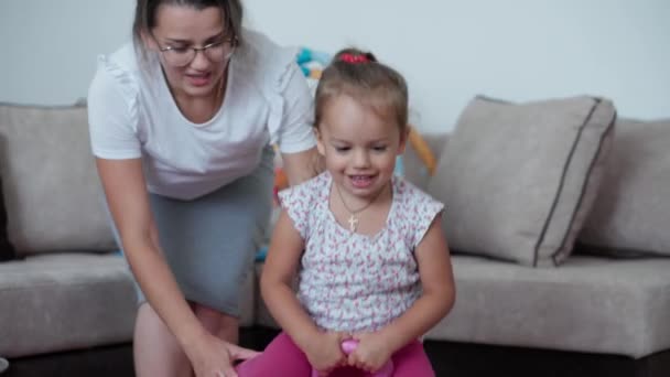 La infancia, los juegos, la educación y el concepto de desarrollo - niña pequeña saltar en el fitball con la mamá linda joven en el fondo hermana recién nacida se encuentra en la estera en la sala de juegos para niños. Los hermanos se divierten en casa en cuarentena — Vídeos de Stock