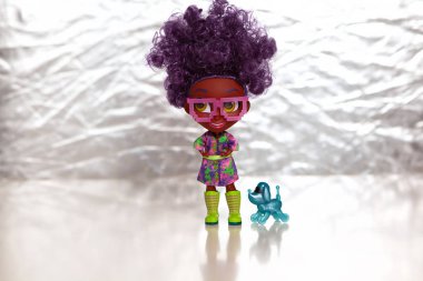 Moskova, Rusya - 18 Nisan 2019: Doll surprise Hairdorables. Seri 1. Kalın kıvırcık saçlı gözlüklü koyu tenli bebek. Evcil köpek robotuyla. Parlak gümüş arka plan.