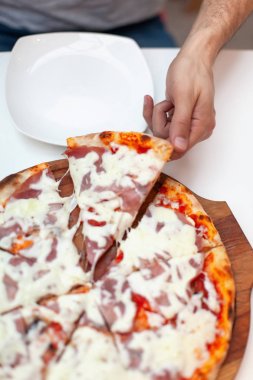 Adam lezzetli İtalyan pizzailk dilim aldı. Çerçeve el ahşap tahta üzerinde jambon ile sıcak pizza dilim alarak, beyaz masa üzerinde beyaz servis tabağı. Pizza zamanı. Üstte görüntü.