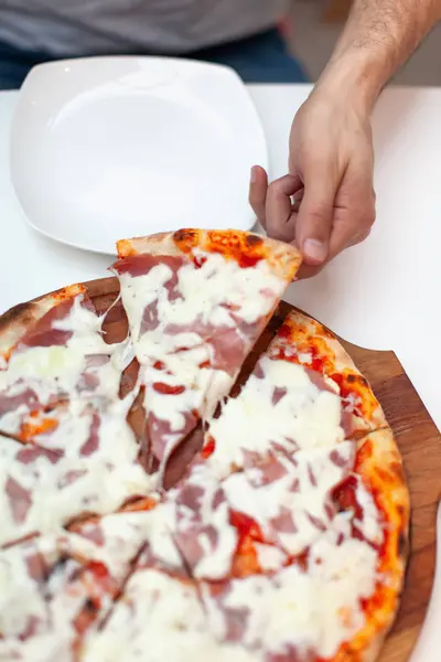 L'uomo ha preso la prima fetta di deliziosa pizza italiana. In cornice presa a mano fetta di pizza calda con prosciutto su tavola di legno, piatto da portata bianco su tavolo bianco. E 'ora della pizza. Vista dall'alto . — Foto Stock