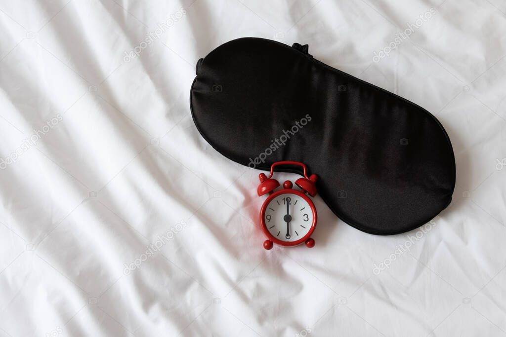 Silk black sleep mask, red clock, showing 6 o'clock on white rumpled sheets. Top view, flat lay. Horizontal. Concept of rest, awakening, sleep, good morning. For social media, blog. Minimal style.