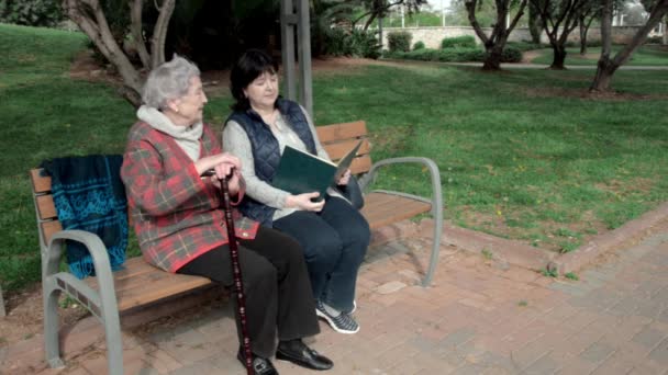 キャラーの仲間は 都市公園の木製のベンチに座っている老婦人のための本を読みます 先輩女性は注意深く耳を傾け うなずく 彼女はこの介護者が好きです — ストック動画