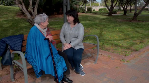 成熟的女性护理员和一位年长的女性坐在城市公园的长椅上交谈 照顾者把倒下的蓝色披肩放在她的病房里 — 图库视频影像
