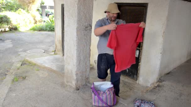 ホームレスの男性は ゴミ部屋の中古着の中に赤いTシャツを見つけたことをとても喜んでいます — ストック動画