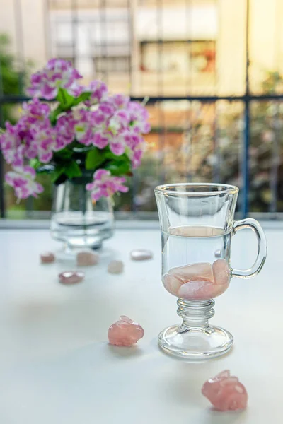 In Irish coffee mug preparing drinkable gem elixir. Burnished rose quartz stones are placed into distilled water. Two stone rabbits sit nearby it. Vase with flowers is seen on the balcony table.