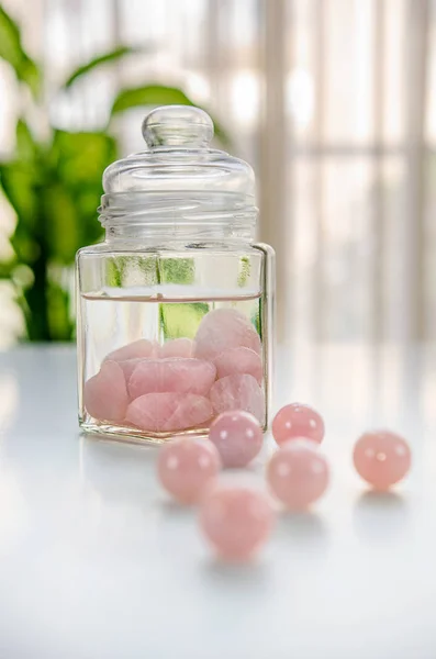 Burnished rose quartz stones are put inside of a glass jar. The drinking water is being charged with the healing energy of crystals. Six  beads are in the foreground out of focus.