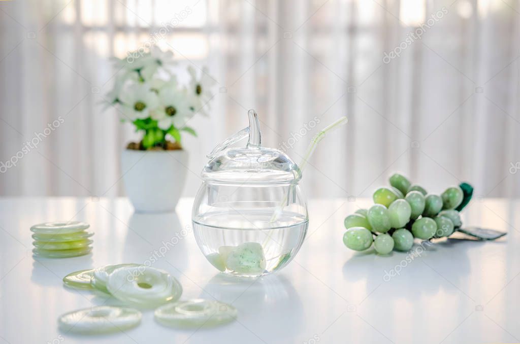 Polished small jade stones are located inside of a glass jar with crystalline water. It is making crystal charged water at home. There are donuts carved grapes and white flowers on the table.
