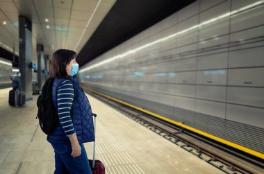 Metro istasyonunda tren beklerken mavi koruyucu maske takan orta yaşlı bir kadının yan portresi. Bagajı, siyah bir sırt çantası ve bordo bir bavulu var. Metro platformu neredeyse boş..