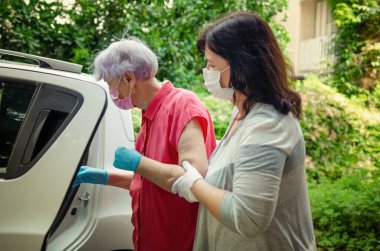Carer helps her elderly female client to get into a  white car to transport for regular medical or any other appointments. Both wear protective masks. clipart