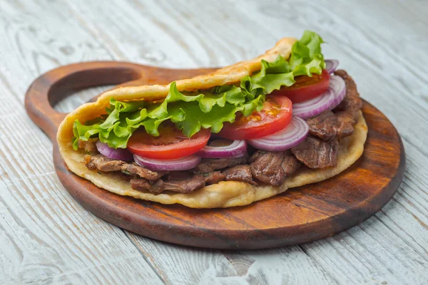 Fresh homemade pita sandwich with roasted meat, tomato, onion and lettuce on wooden board on kitchen table.