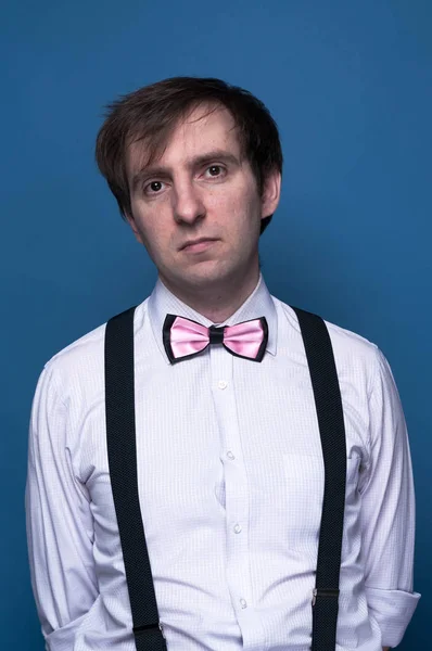 handsome man in shirt, pink bow tie and black suspenders looking at camera on blue background