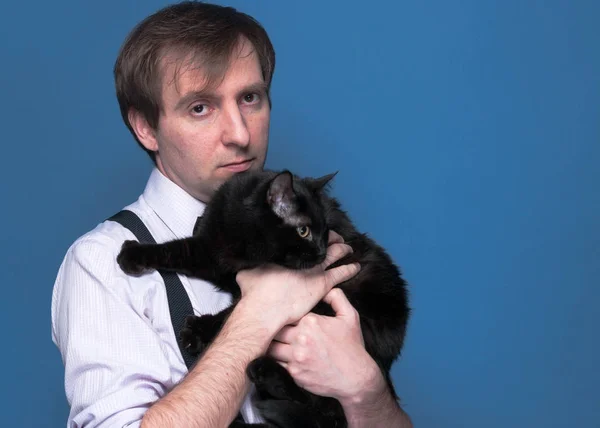 handsome man in shirt and suspender holding and hugging black funny cat on blue background with copy space