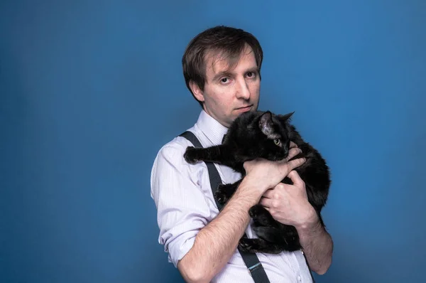 handsome man in shirt and suspender holding and hugging black funny cat on blue background