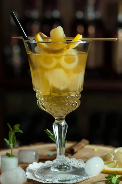 yellow alcoholic drink in glass with black drinking straw, lemon slices and herbs on dark background in bar