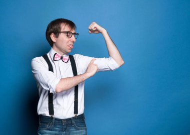 serious handsome man in shirt, suspender, pink bow tie and glasses standing and holding smartphone on blue background with copy space 