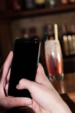 barda ahşap tezgahüzerinde buz ve saman ile kırmızı alkollü içecek önünde boş skreen ile akıllı telefon tutan adam kırpılmış görünümü