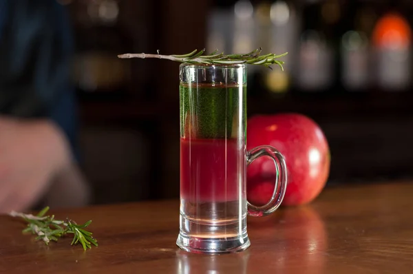 selective focus of shot glass with colorful alcohol drink and rosemary near red apple on wooden counter in bar