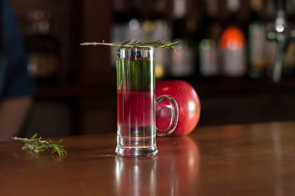 selective focus of shot glass with multicolored alcohol drink and rosemary on near red apple on wooden table in bar