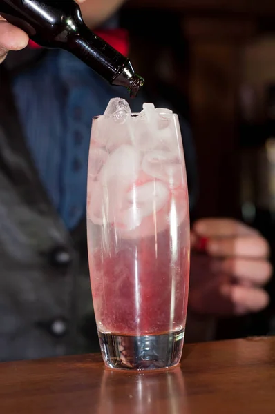 バーの木製のテーブルの上にアルコール飲料と氷でボトルからガラスに赤のシロップを注ぐバーテンダーのクロップビュー — ストック写真