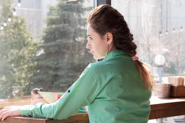 back view of woman with french braid in green shirt sitting at wooden table and looking at window in coffee shop