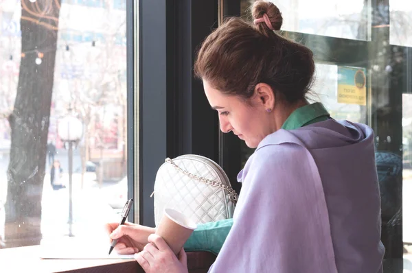 side view of woman with bun in purple scarf sitting at wooden table writing in notebook with pen and holding paper cup in coffee shop