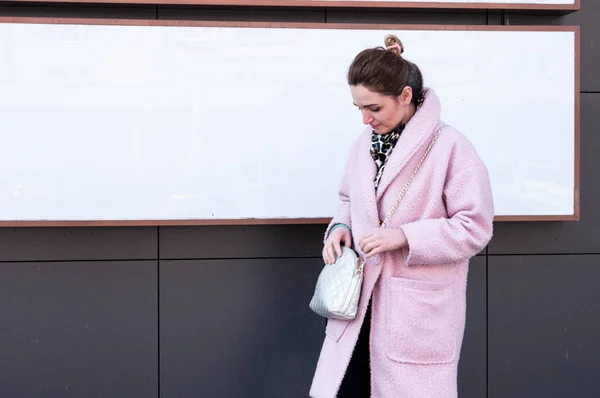暗い大胆に大きな空のボードの近くに立って 路上でバッグを見てピンクのコートで魅力的な自信のある女性 — ストック写真